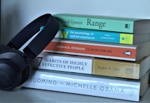 A stack of five books with diverse topics ranging from psychology to personal development sits on a white surface. Next to the books, a black set of JBL headphones is positioned, suggesting a relationship between reading and listening.