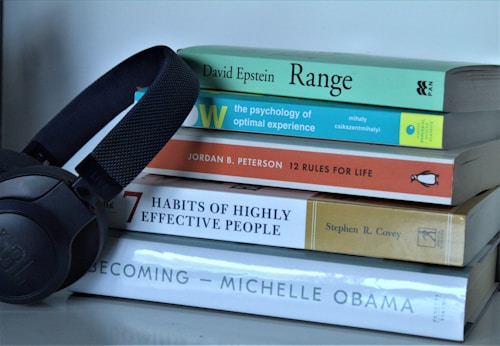 A stack of five books with diverse topics ranging from psychology to personal development sits on a white surface. Next to the books, a black set of JBL headphones is positioned, suggesting a relationship between reading and listening.