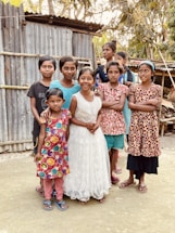 A group of children stands together outdoors in front of a corrugated metal structure. They wear colorful dresses, and one girl is in a white dress. The setting appears rural with trees in the background.