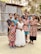 A group of children stands together outdoors in front of a corrugated metal structure. They wear colorful dresses, and one girl is in a white dress. The setting appears rural with trees in the background.