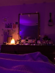 The image depicts a cozy bedroom setup with a dresser topped with various items, such as a salt lamp that's lit, bottles, and decorative objects. The room is dimly lit with purple string lights adding a warm glow. A mirror above the dresser reflects some of the room, and there are decorative elements like pictures and a ukulele hanging on the wall.