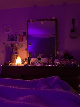 The image depicts a cozy bedroom setup with a dresser topped with various items, such as a salt lamp that's lit, bottles, and decorative objects. The room is dimly lit with purple string lights adding a warm glow. A mirror above the dresser reflects some of the room, and there are decorative elements like pictures and a ukulele hanging on the wall.
