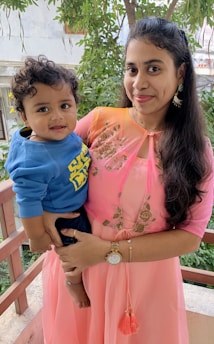 A woman is holding a smiling child in a cozy outdoor setting surrounded by greenery. She is wearing a pink traditional dress while the child is dressed in a blue sweatshirt. Both appear happy and content.