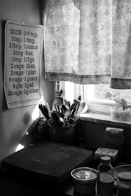 A kitchen scene featuring a wall chart with cooking measurements, a floral-patterned curtain partially covering a window, and several kitchen items like utensils in a holder, a bottle, and containers on a countertop.