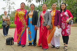 A warm group photo of diverse women smiling together, wearing pink scarves in a sunlit garden.