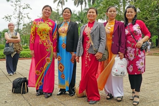 A group of six women standing together outdoors, with five of them wearing colorful traditional dresses featuring floral and geometric patterns. They are posing happily for the camera. The background includes greenery and trees, indicating a pleasant park setting.