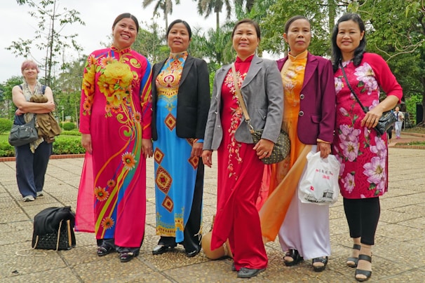A group of six women standing together outdoors, with five of them wearing colorful traditional dresses featuring floral and geometric patterns. They are posing happily for the camera. The background includes greenery and trees, indicating a pleasant park setting.