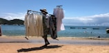 A person walks along a sandy beach, carrying a clothes rack filled with light, flowing garments. The scene features a calm sea with small boats floating in the water and a few people swimming, set against a backdrop of hills and a partly cloudy sky.