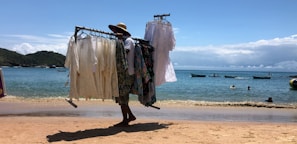 A person walks along a sandy beach, carrying a clothes rack filled with light, flowing garments. The scene features a calm sea with small boats floating in the water and a few people swimming, set against a backdrop of hills and a partly cloudy sky.