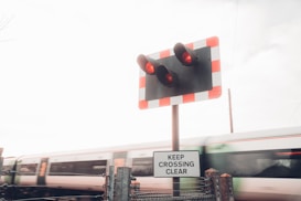 A railway crossing scene features blurred motion of a passing train in the background. There is a prominent red and white striped railroad crossing sign with two black warning lights. A sign below reads 'KEEP CROSSING CLEAR'. The image conveys a sense of movement with the train zooming past the stationary warning signs.