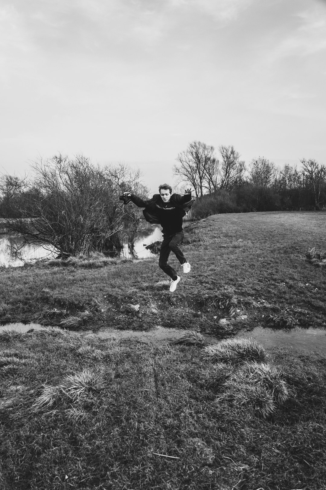 grayscale photo of man in black jacket and pants jumping on grass field