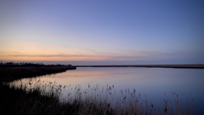 A serene landscape shot at dusk with a calm lake reflecting the pastel colors of the sky, framed in a wide horizontal format.