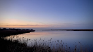 A serene landscape shot at dusk with a calm lake reflecting the pastel colors of the sky, framed in a wide horizontal format.