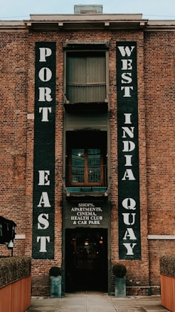 A brick building facade featuring large vertical signs with bold white letters. The words 'Port East' and 'West India Quay' are prominently displayed. Below, a smaller sign indicates the presence of shops, apartments, a cinema, a health club, and a car park.