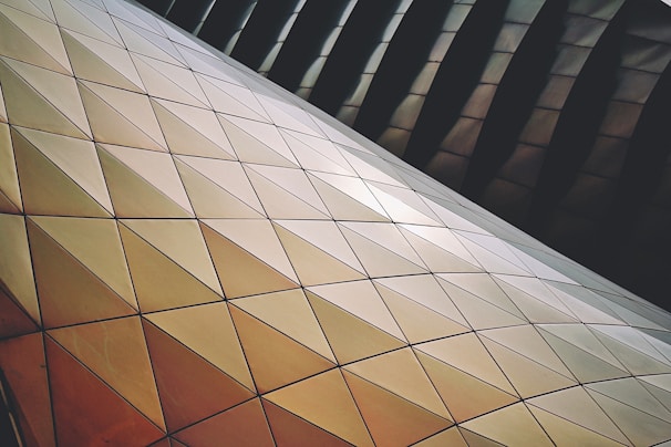 A series of interlocking geometric panels with triangular shapes, featuring a gradient of warm tones. The panels form a complex, futuristic structure with a glossy surface, reflecting light and creating a textured pattern.