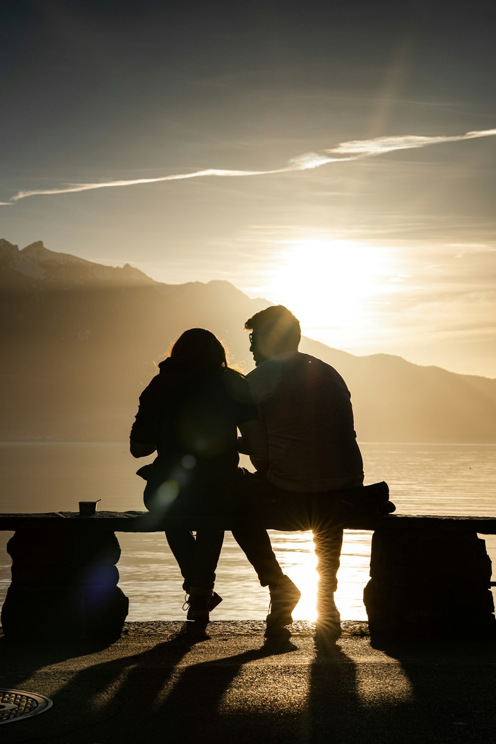 silhueta do homem e da mulher sentados no banco perto do corpo de água durante o pôr do sol