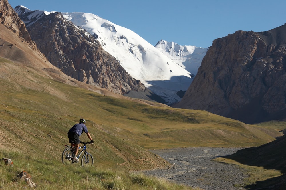 man in blue jacket riding bicycle on green grass field near snow covered mountain during daytime