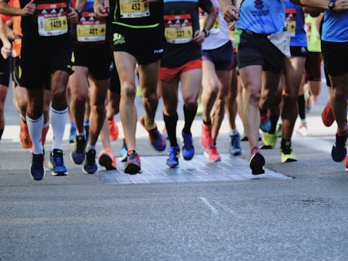 A group of runners is captured in motion during a marathon or race. The focus is on their legs and feet, showing a variety of running shoes and athletic wear. The ground is a paved road, and the runners display different race bibs on their attire.
