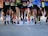 A group of runners is captured in motion during a marathon or race. The focus is on their legs and feet, showing a variety of running shoes and athletic wear. The ground is a paved road, and the runners display different race bibs on their attire.