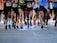 A group of runners is captured in motion during a marathon or race. The focus is on their legs and feet, showing a variety of running shoes and athletic wear. The ground is a paved road, and the runners display different race bibs on their attire.