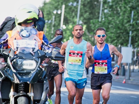A group of runners participating in a road race, accompanied by a motorcycle with a photographer capturing the event. The runners are wearing athletic gear and race bibs, appearing focused and determined as they progress through the course. The background includes blurred greenery, creating a sense of motion.