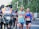 A group of runners participating in a road race, accompanied by a motorcycle with a photographer capturing the event. The runners are wearing athletic gear and race bibs, appearing focused and determined as they progress through the course. The background includes blurred greenery, creating a sense of motion.