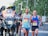 A group of runners participating in a road race, accompanied by a motorcycle with a photographer capturing the event. The runners are wearing athletic gear and race bibs, appearing focused and determined as they progress through the course. The background includes blurred greenery, creating a sense of motion.
