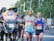 A group of runners participating in a road race, accompanied by a motorcycle with a photographer capturing the event. The runners are wearing athletic gear and race bibs, appearing focused and determined as they progress through the course. The background includes blurred greenery, creating a sense of motion.