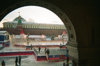 A festive winter scene with an outdoor ice skating rink surrounded by decorations, including red and white tents and holiday lights. The rink is situated in front of a large historic building with a Russian architectural style, topped with a green dome and a flag. Snow is lightly covering the ground, and a few people are walking or standing nearby, some appearing to enjoy the ice skating.