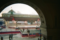 A festive winter scene with an outdoor ice skating rink surrounded by decorations, including red and white tents and holiday lights. The rink is situated in front of a large historic building with a Russian architectural style, topped with a green dome and a flag. Snow is lightly covering the ground, and a few people are walking or standing nearby, some appearing to enjoy the ice skating.