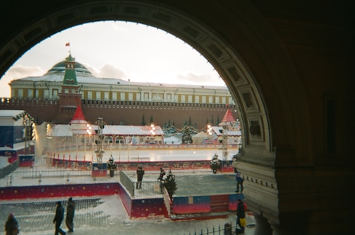 A festive winter scene with an outdoor ice skating rink surrounded by decorations, including red and white tents and holiday lights. The rink is situated in front of a large historic building with a Russian architectural style, topped with a green dome and a flag. Snow is lightly covering the ground, and a few people are walking or standing nearby, some appearing to enjoy the ice skating.