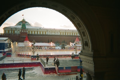 A festive winter scene with an outdoor ice skating rink surrounded by decorations, including red and white tents and holiday lights. The rink is situated in front of a large historic building with a Russian architectural style, topped with a green dome and a flag. Snow is lightly covering the ground, and a few people are walking or standing nearby, some appearing to enjoy the ice skating.