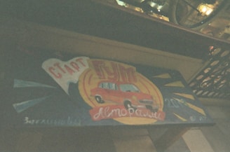 A vintage-style sign featuring a red car against a backdrop with a blue and yellow color scheme. The sign includes Cyrillic text and conveys a retro automotive theme. It seems to be set against a wood or metal panel, possibly part of a storefront or display.