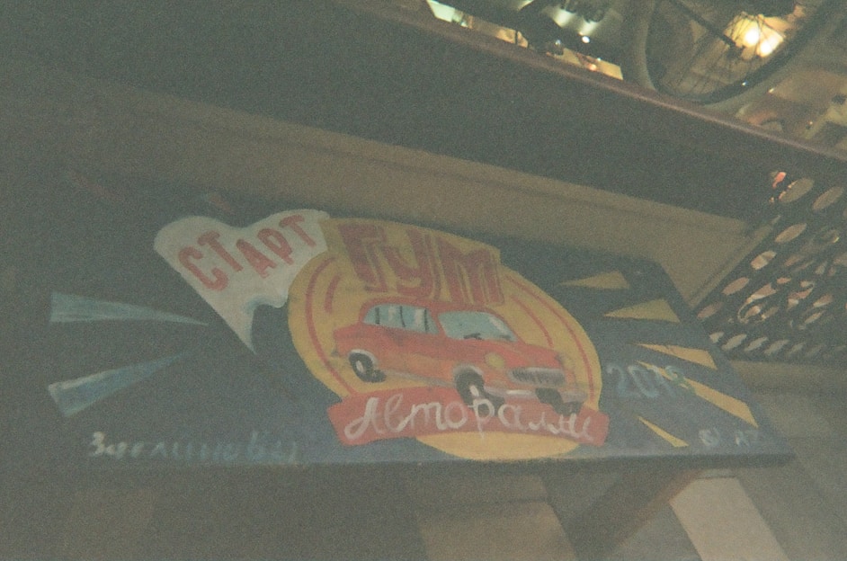 A vintage-style sign featuring a red car against a backdrop with a blue and yellow color scheme. The sign includes Cyrillic text and conveys a retro automotive theme. It seems to be set against a wood or metal panel, possibly part of a storefront or display.