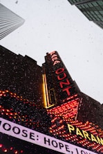 A tall building with a bright marquee sign displaying 'PARA' and 'OUT' in red neon lights. The building is set against a gray sky with falling snowflakes, and other skyscrapers are visible in the background. The marquee also features other illuminated text and decorative lights in red and yellow.