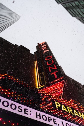 A tall building with a bright marquee sign displaying 'PARA' and 'OUT' in red neon lights. The building is set against a gray sky with falling snowflakes, and other skyscrapers are visible in the background. The marquee also features other illuminated text and decorative lights in red and yellow.