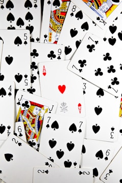 A scattered pile of playing cards, primarily focusing on spades and clubs suits, featuring a prominent ace of hearts in the center. The face cards include a Jack of Clubs and a King of Hearts with elaborate designs. The cards are arranged in a seemingly random manner, creating a sense of movement and chaos.
