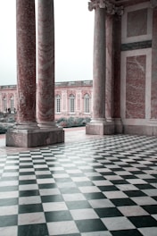 Large marble columns with intricate detailing stand on a black and white checkered floor, leading to an open area with a historic building featuring multiple arched windows and elegant stonework.