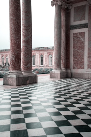 Large marble columns with intricate detailing stand on a black and white checkered floor, leading to an open area with a historic building featuring multiple arched windows and elegant stonework.