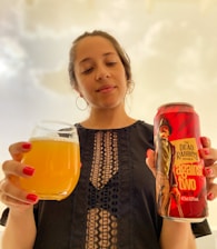 A person holding a glass of orange-yellow drink in one hand and a can with a red label featuring 'The Dead Rabbits' and 'against two' in the other. The person is wearing a black top with intricate lace patterns. The background is softly lit, creating a warm and inviting atmosphere.