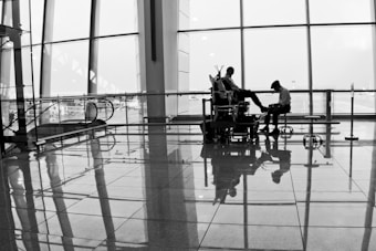 A monochrome image of an airport terminal showing a man sitting in a chair, receiving a shoe shine from another person. The scene captures their reflections on the shiny floor, with large glass windows in the background displaying an overcast sky and the silhouette of the airport infrastructure.
