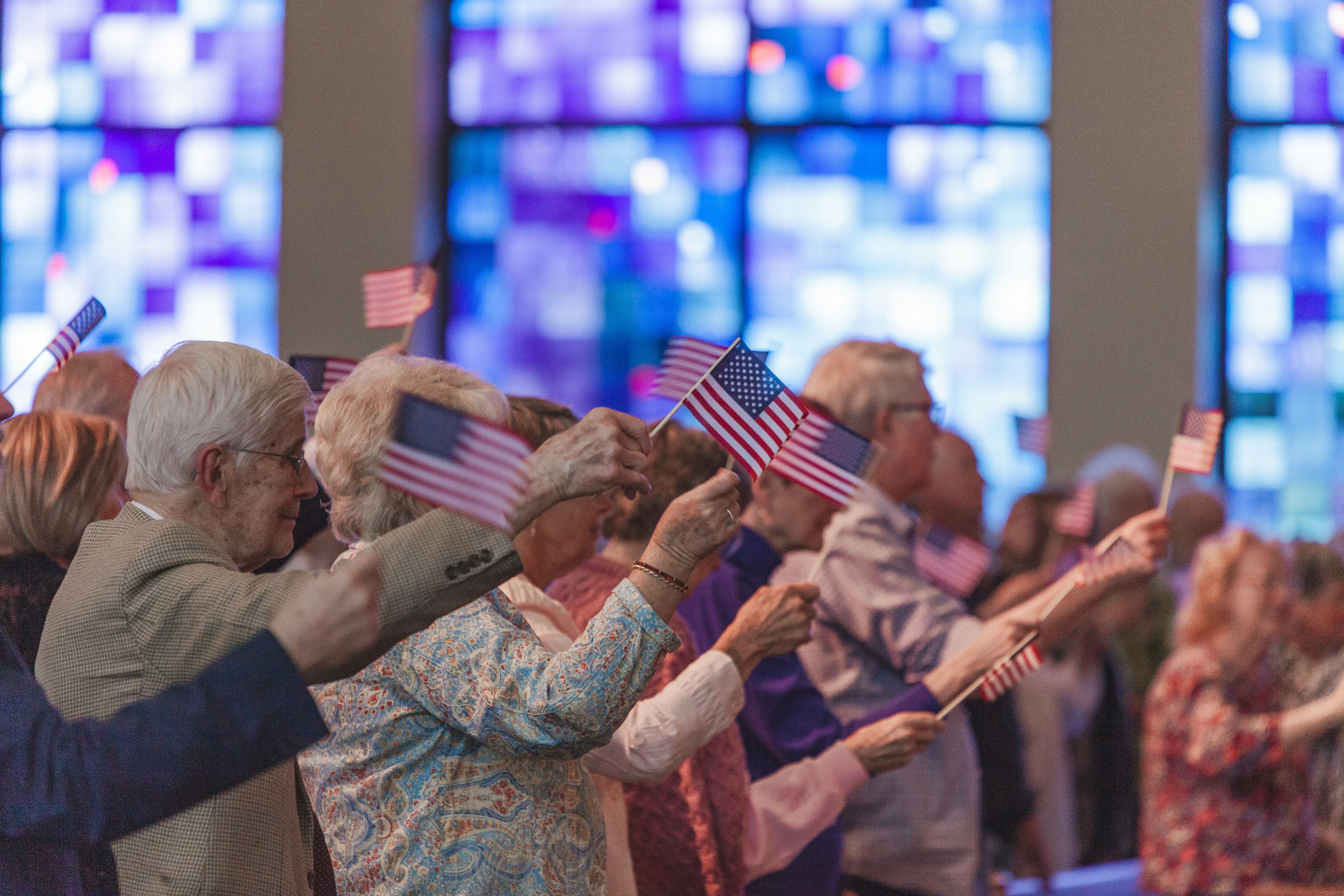 Elderly men and women waving tiny American flags at an indoor gathering.