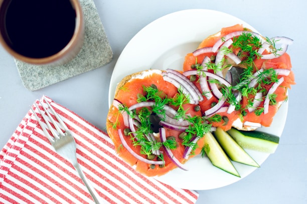 A plate with bagels topped with smoked salmon, red onion slices, dill, and capers is displayed alongside sliced cucumbers. A red and white striped napkin with a fork on it is placed nearby. A cup of coffee sits on a stone coaster.
