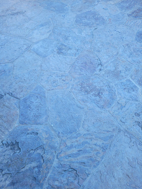 A close-up of a textured stone surface with irregular, polygonal shapes and a slightly rough appearance. The stones are primarily in shades of blue and gray, displaying natural variations in color and pattern.