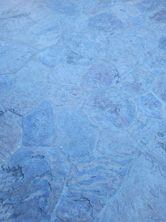 A close-up of a textured stone surface with irregular, polygonal shapes and a slightly rough appearance. The stones are primarily in shades of blue and gray, displaying natural variations in color and pattern.