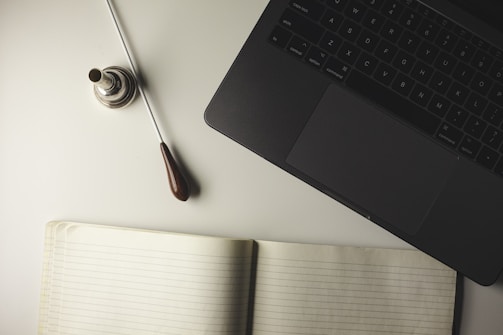 A neat arrangement including a closed laptop, an open lined notebook, and a conductor's baton with a silver base. The items are placed on a light-colored surface, suggesting a workspace or study setting.