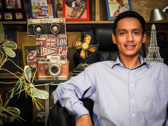 A young man with braces is sitting in a black leather chair, wearing a light blue shirt. Behind him are various decorative items including a vintage camera, a gold retro fan, a small Eiffel Tower statue, and framed pictures on a shelf. There are also some green plants on the left side of the image.