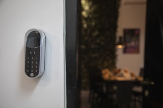 A smart digital lock with a keypad is mounted on a white wall. In the background, there is a blurred view of a dining area with a table and chairs, and some wall art is partially visible.