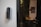 A smart digital lock with a keypad is mounted on a white wall. In the background, there is a blurred view of a dining area with a table and chairs, and some wall art is partially visible.