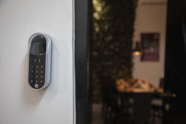 A smart digital lock with a keypad is mounted on a white wall. In the background, there is a blurred view of a dining area with a table and chairs, and some wall art is partially visible.